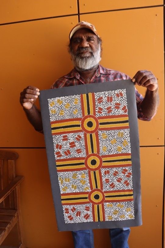 Indigenous man holding a colorful dot-painted artwork, orange wall background.