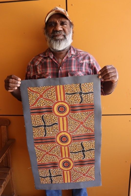 Man holding Aboriginal artwork. He has gray beard, wears cap and plaid shirt, stands against orange wall.