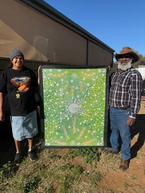Two people holding a green and white dot painting outdoors, under a sunny sky.