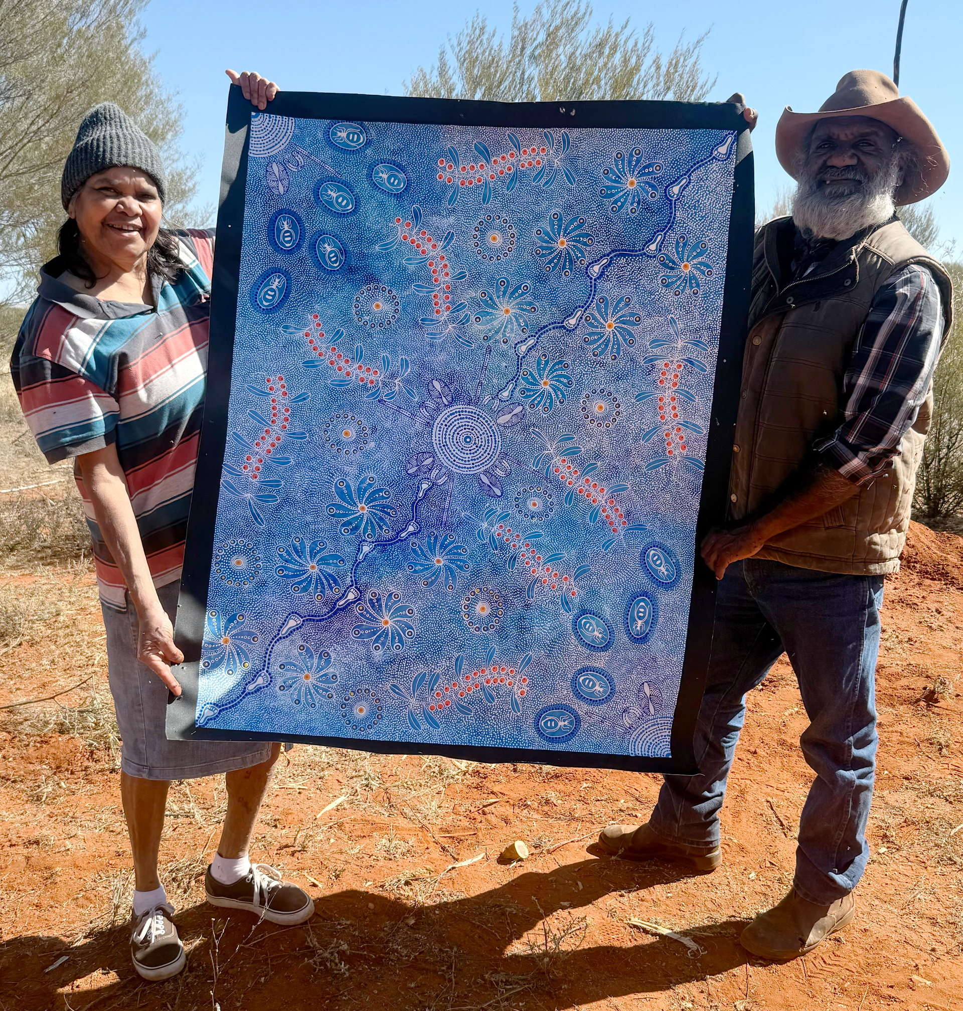 Two people hold up a large abstract painting with blue and orange details, in an outdoor setting.