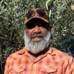 Man with gray beard wearing a hat and sunglasses, orange plaid shirt, outside in front of foliage.