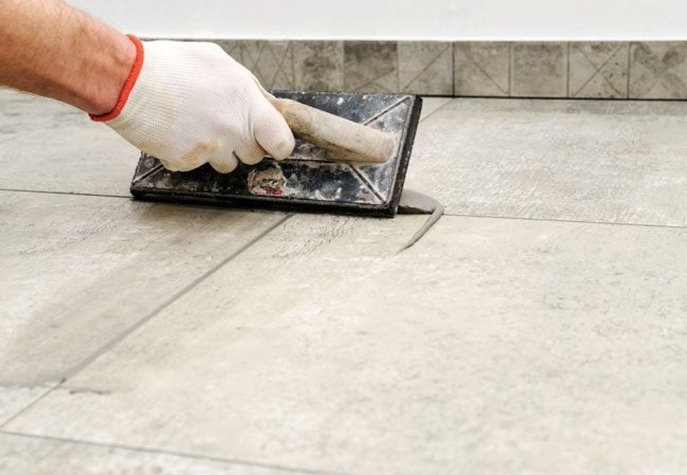 Person using a trowel to apply grout between floor tiles; white glove, gray tile.