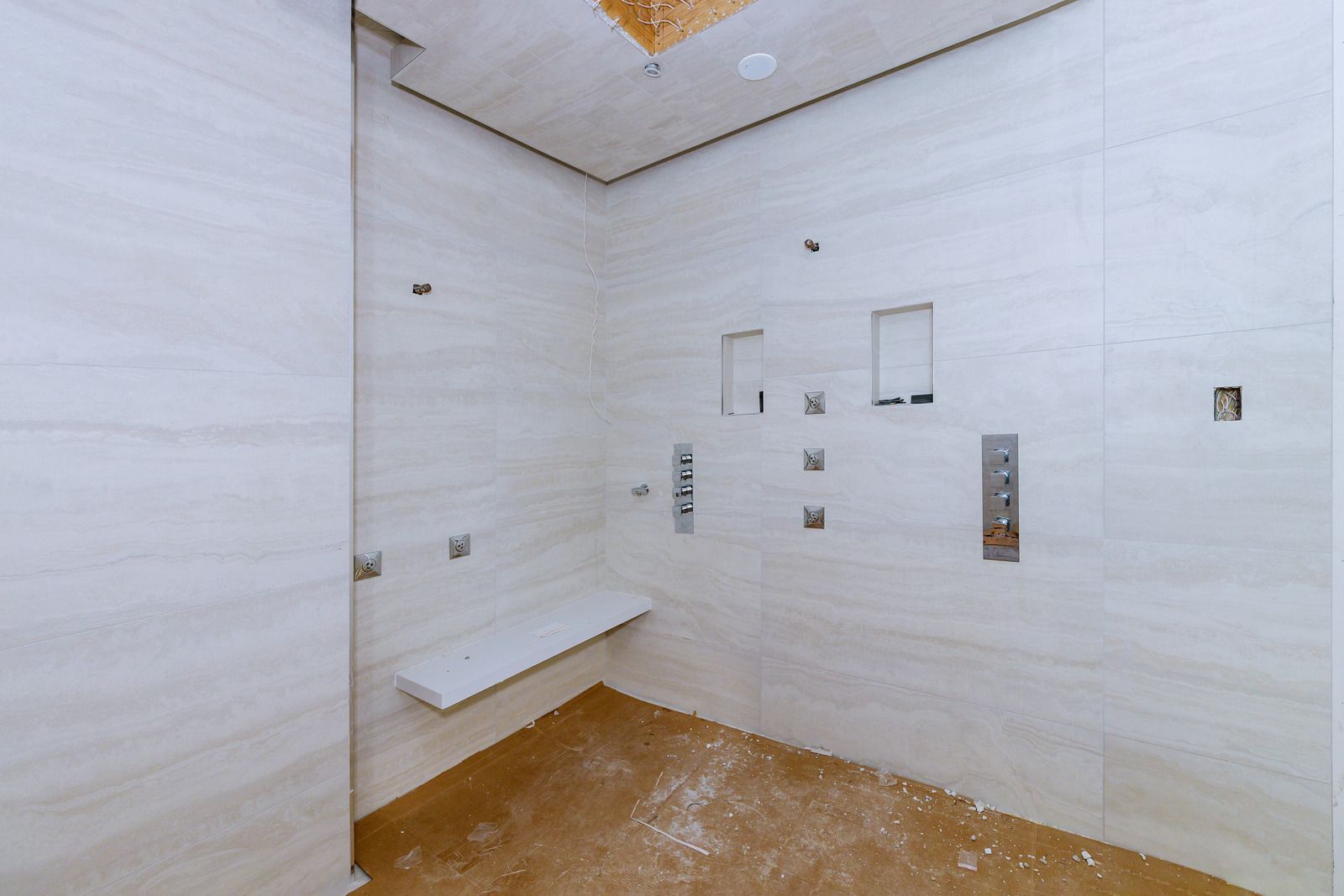 Empty shower with light-colored tile walls, built-in bench, and controls. The floor is covered in debris.