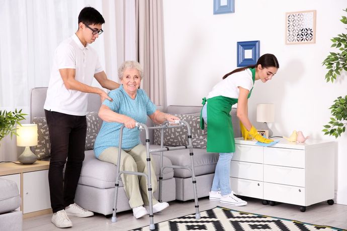 A Man is Helping an Elderly Woman With a Walker in a Living Room — Home Nursing Services In Macquarie, NSW