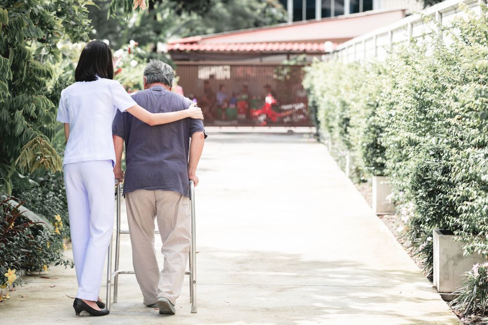 A Nurse Is Helping An Elderly Man Walk With A Walker — Home Nursing Services In Williamtown, NSW