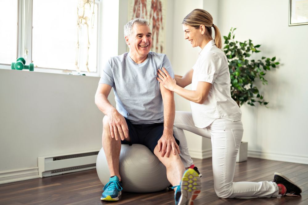 A Man is Talking to a Woman Who is Sitting on a Table — Home Nursing Services In Laurieton, NSW