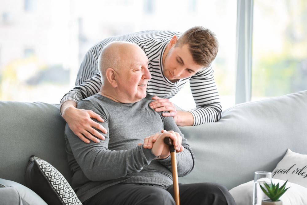 An Elderly Man With A Cane Is Sitting On A Couch With A Young Man — Home Nursing Services In Fennell Bay, NSW