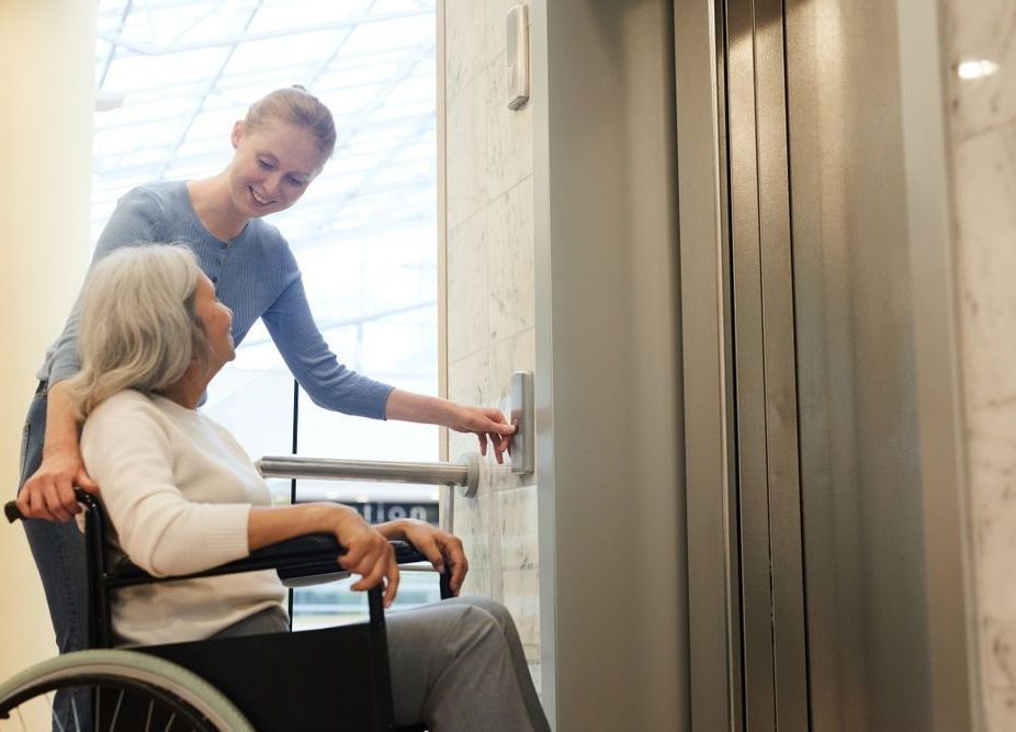 A Woman is Helping an Elderly Woman in a Wheelchair Get Into an Elevator — Home Nursing Services In Wauchope, NSW