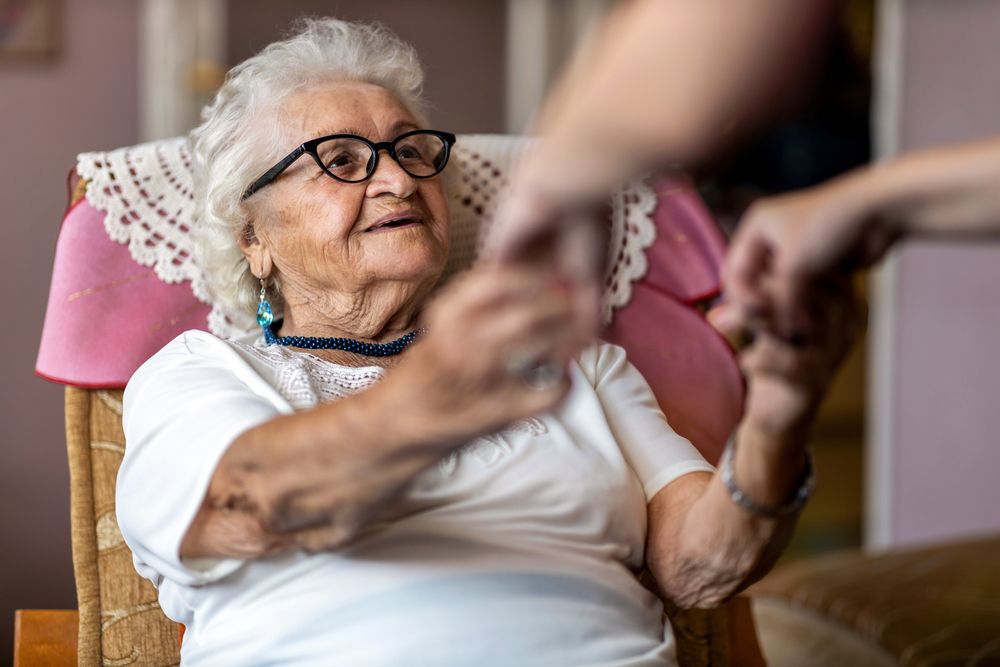 An Elderly Woman Is Sitting In A Chair Holding A Person's Hand — Home Nursing Services In Charlestown, NSW