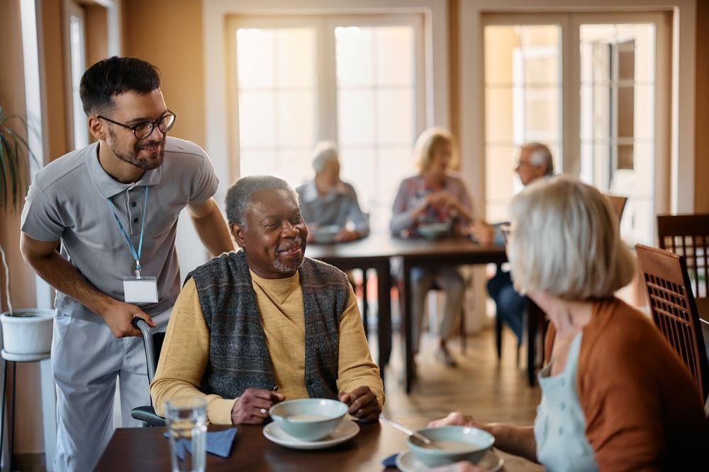 A Man Is Talking To An Elderly Man While Sitting At A Table In A Nursing Home — Home Nursing Services In Medowie, NSW