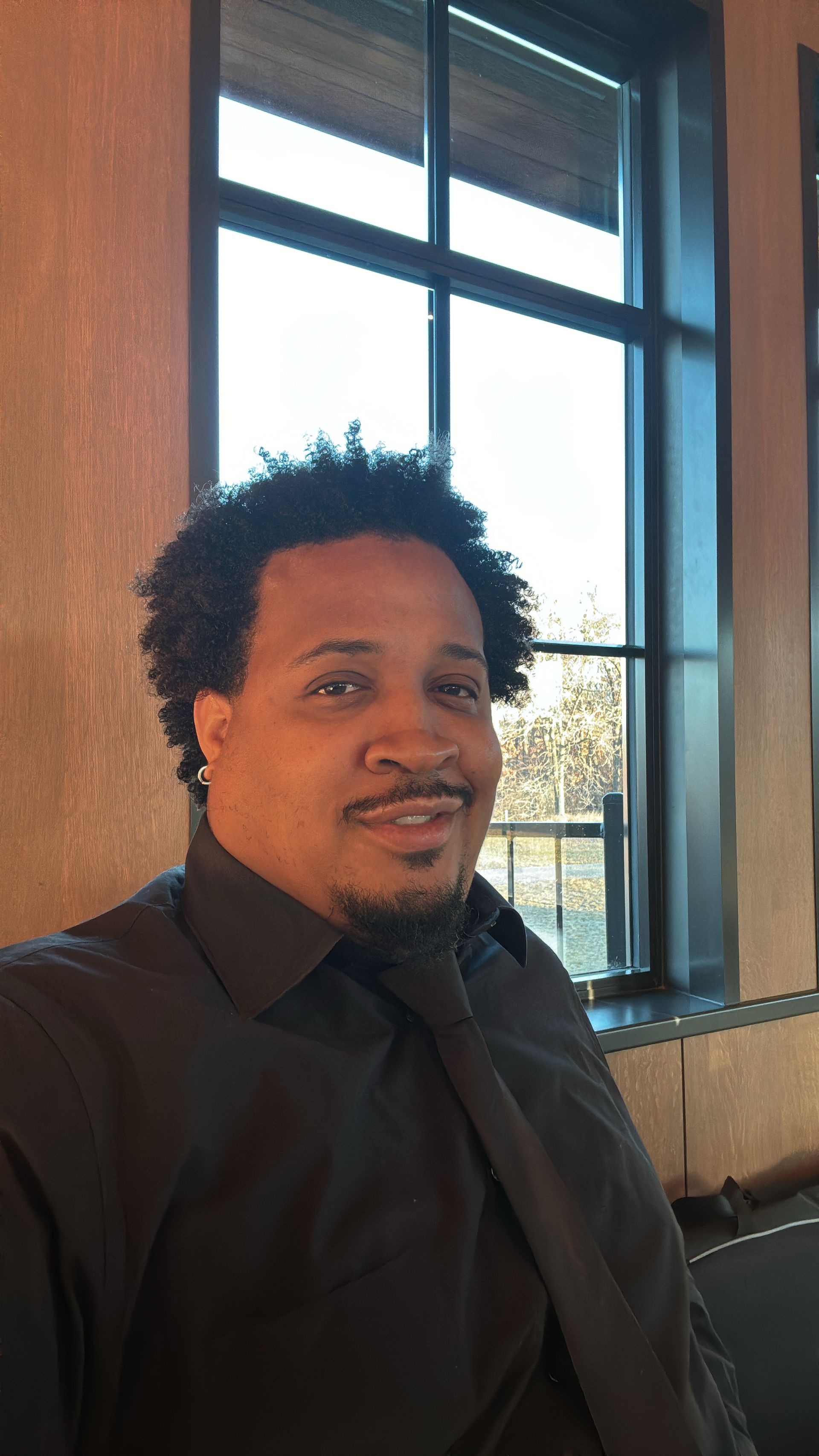 A man in a black shirt and tie is sitting in front of a window.
