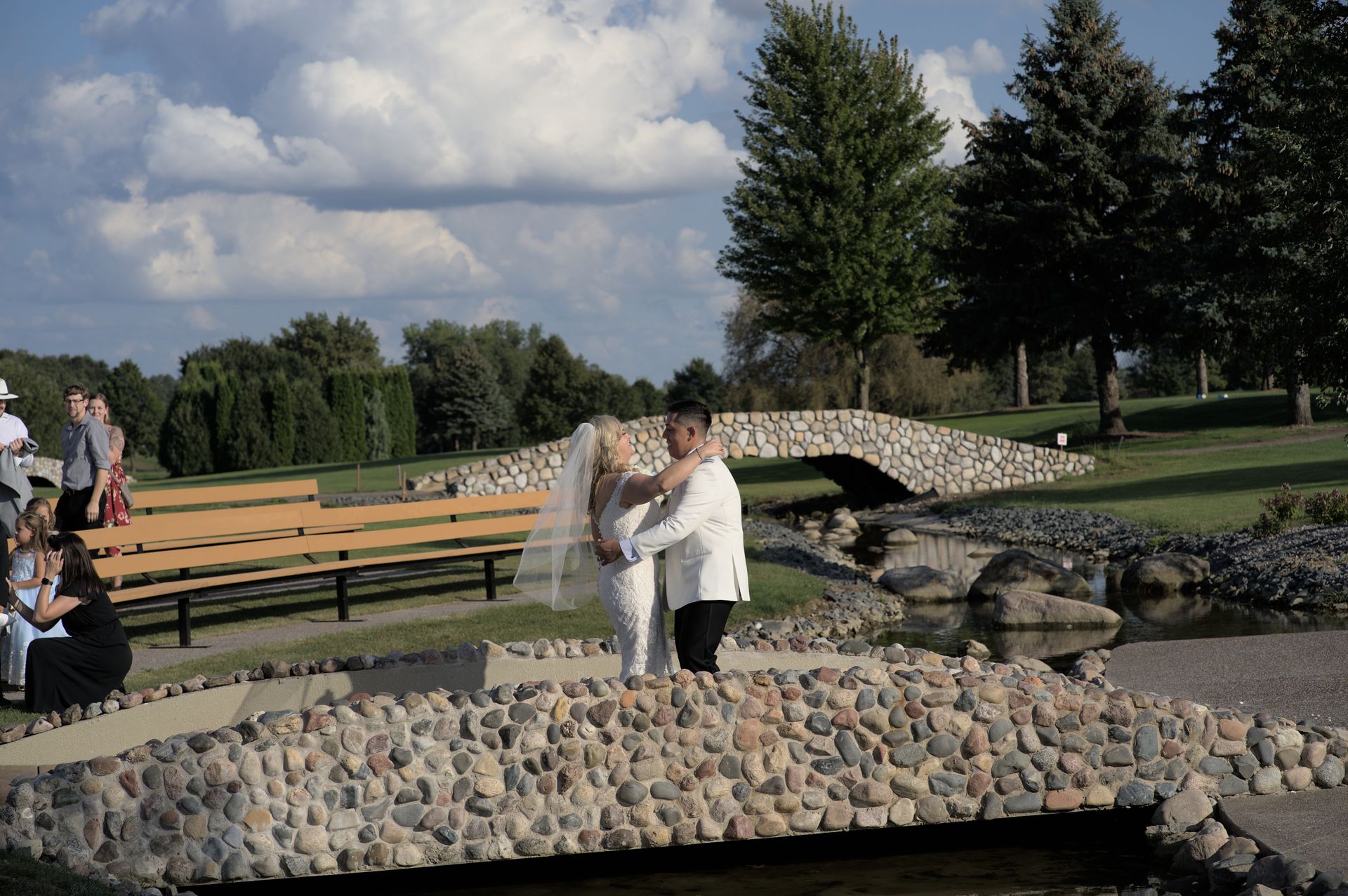 A bride and groom are dancing on a stone bridge in a park.