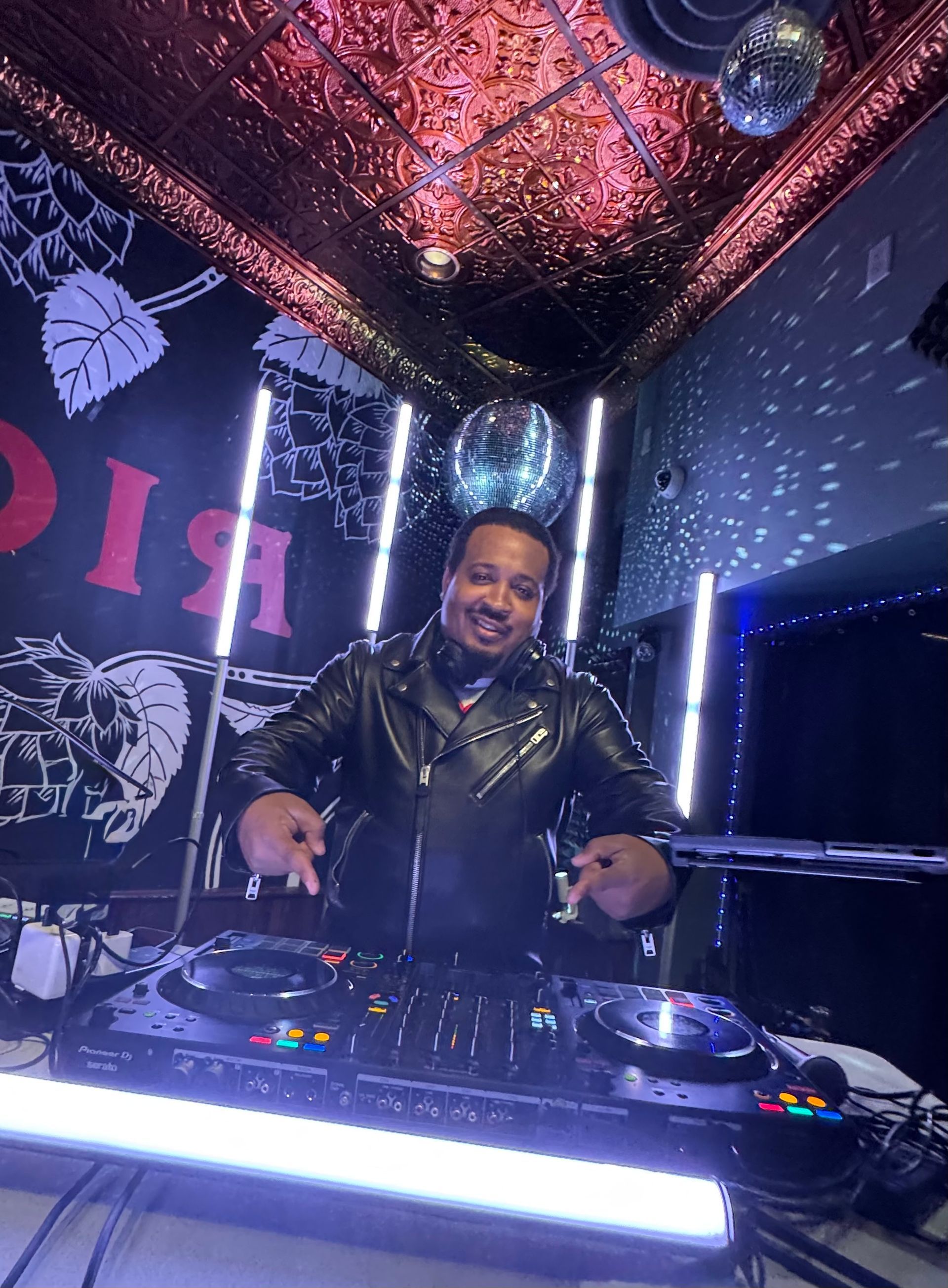 DJ smiling behind a mixer in a neon-lit booth with purple and blue lights