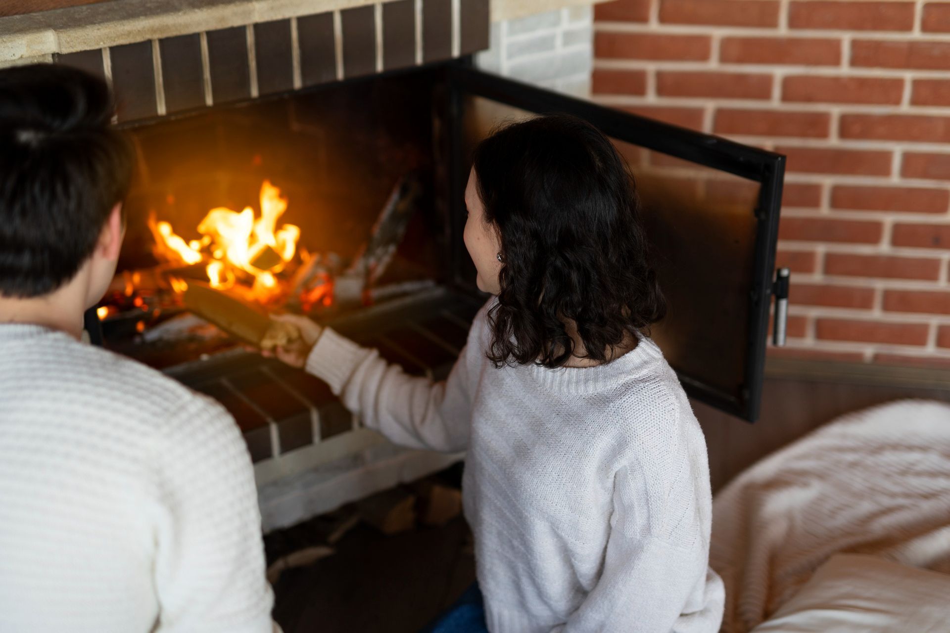 Couple tending a fire in a brick fireplace; woman using a tool on burning logs.