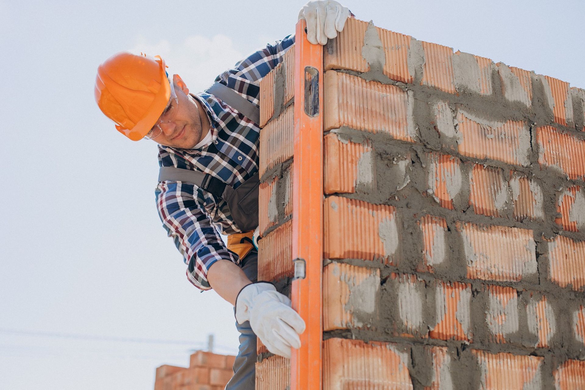 Builder using a level on a brick wall, wearing a hard hat and work gloves outdoors.