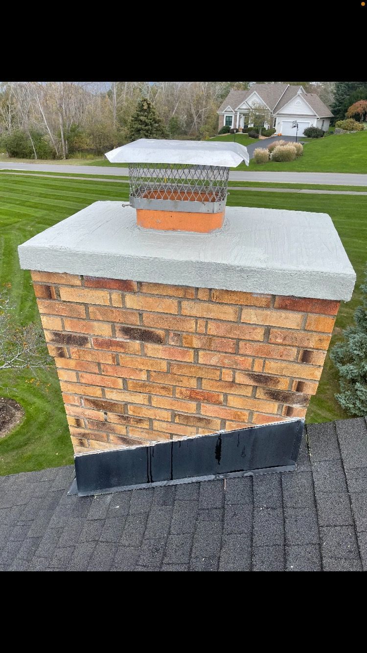 Brick chimney with a metal cap on a rooftop, set against a green lawn and houses.