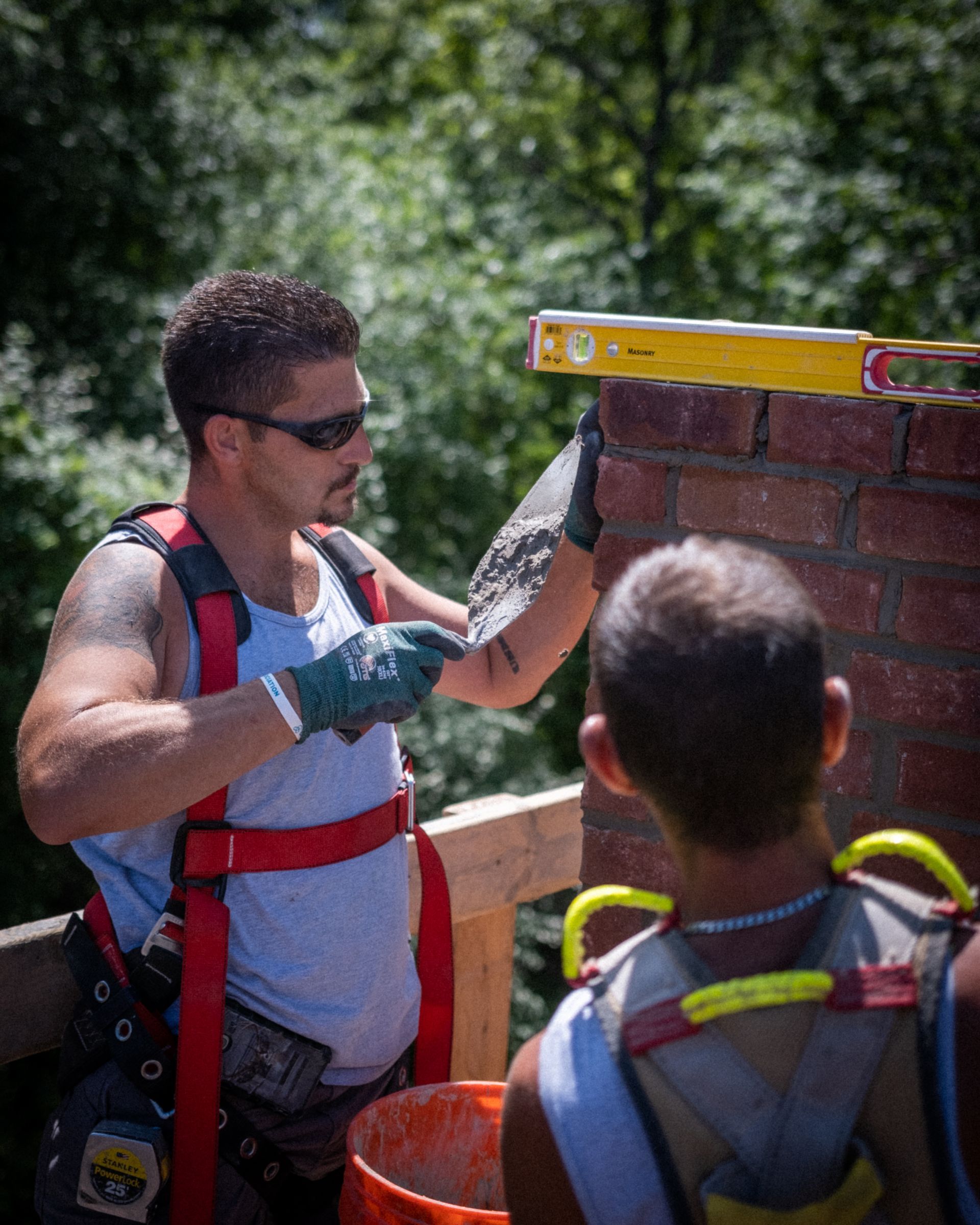 Builder using a level on a brick wall, wearing a hard hat and work gloves outdoors.