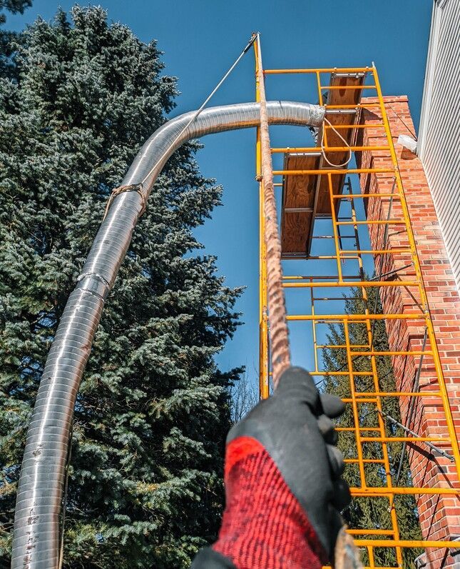 Flexible ductwork being installed on a brick chimney with scaffolding. A gloved hand holds the duct.