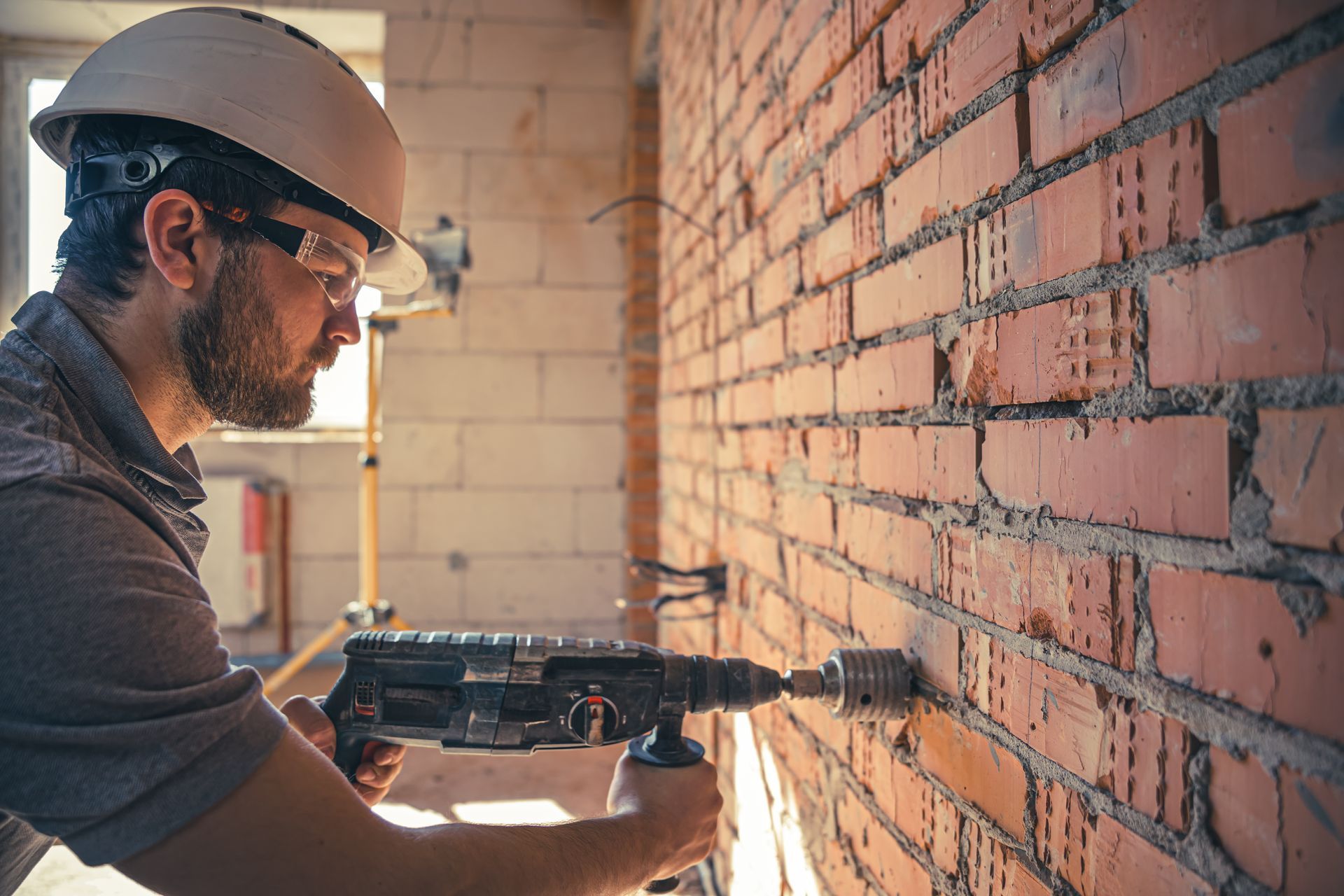 Construction worker drills into a brick wall. He wears safety glasses and a hard hat.