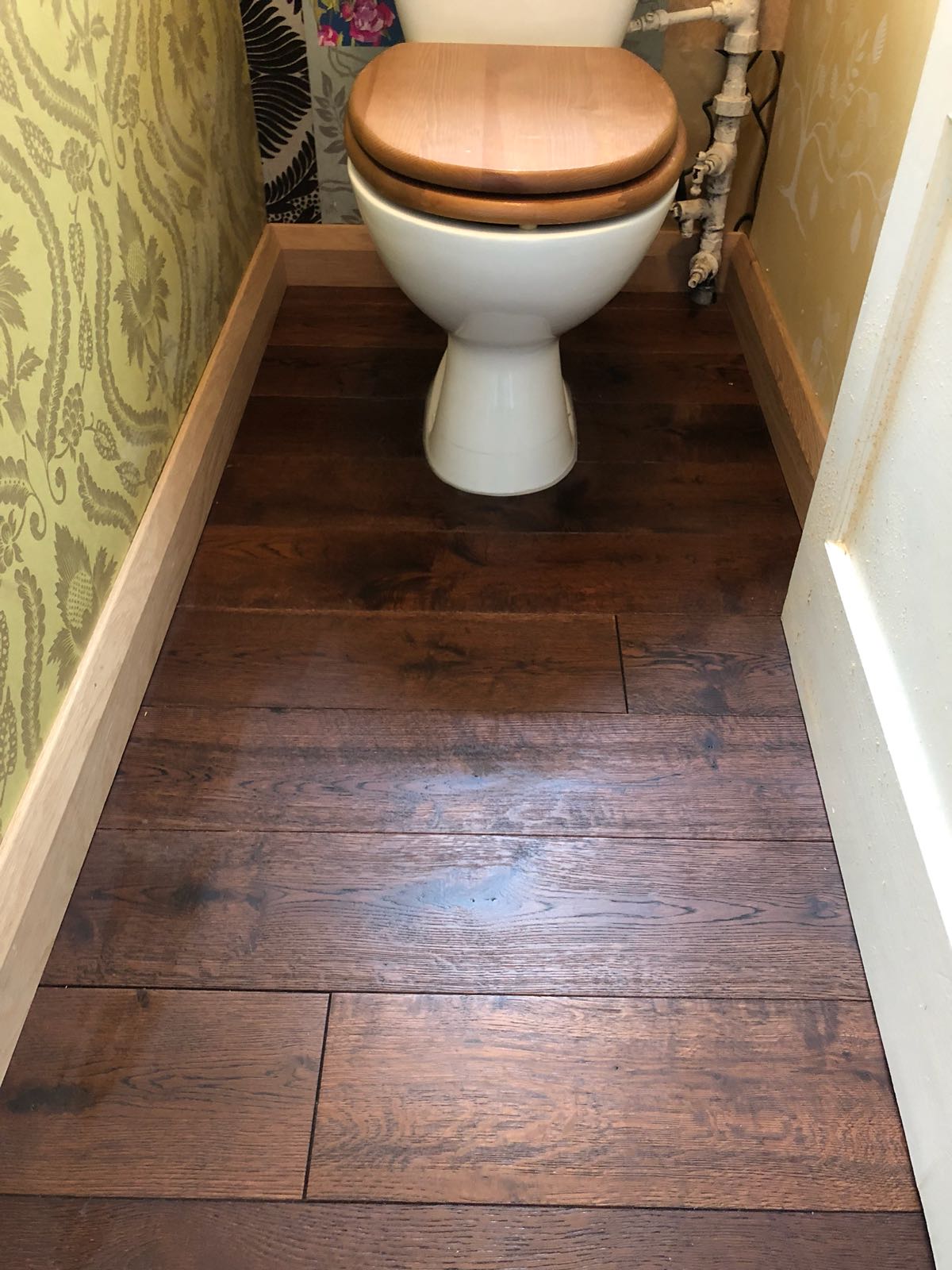 A toilet with a wooden seat is sitting on a wooden floor in a bathroom.