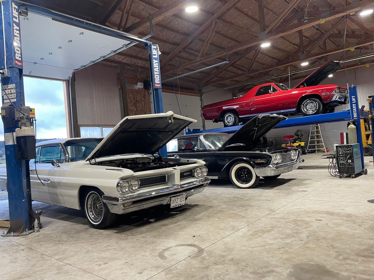 Two old cars are parked in a garage with their hoods open.