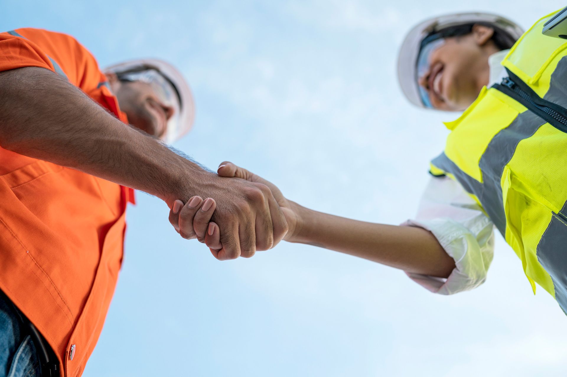 Two construction workers in safety gear shaking hands from low angle.