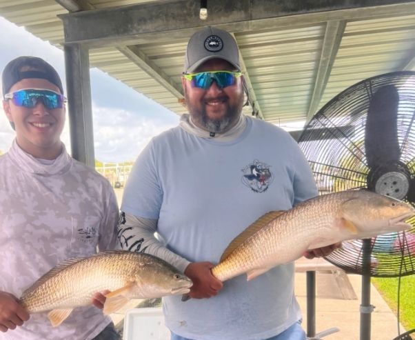 Two men are holding two fish in front of a fan.