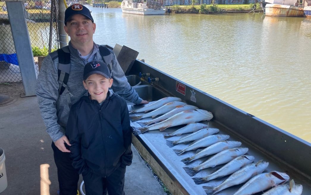 A man and a boy are standing next to a boat full of fish.