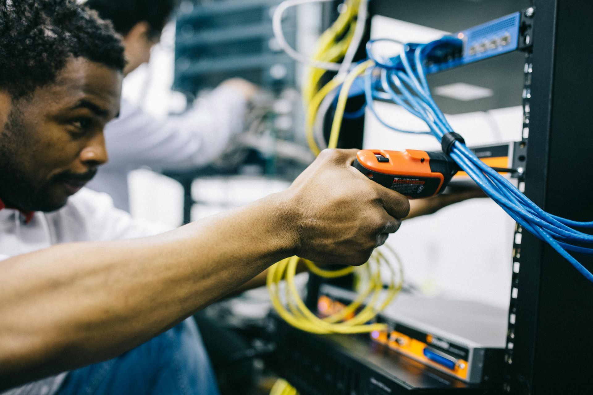 Man working with cables in a server room, holding a tool; another person in background.