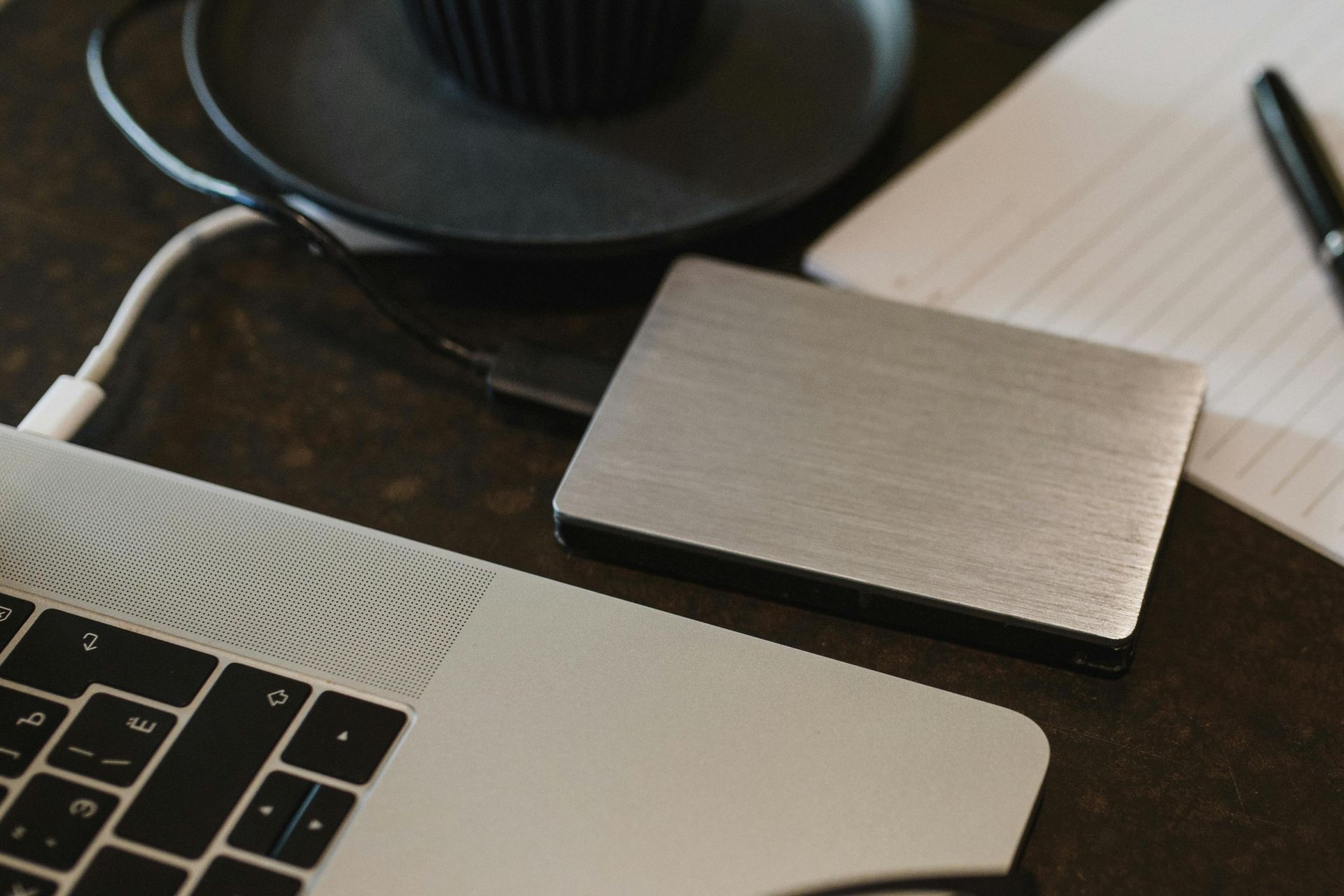 Laptop connected to a silver external hard drive, with a cup and notebook on a table.