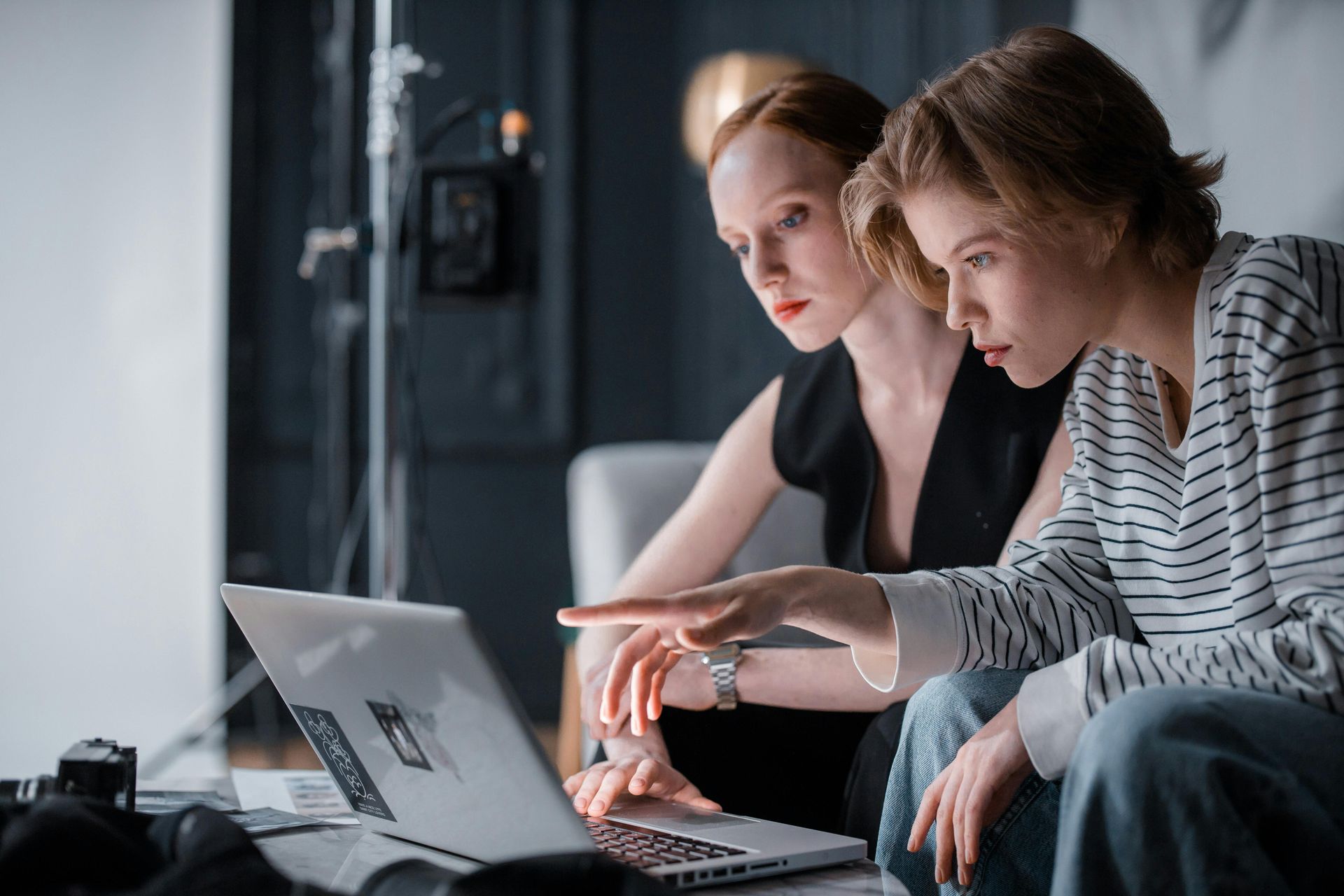 Two women looking at a laptop screen, one points to the screen. Studio setting.