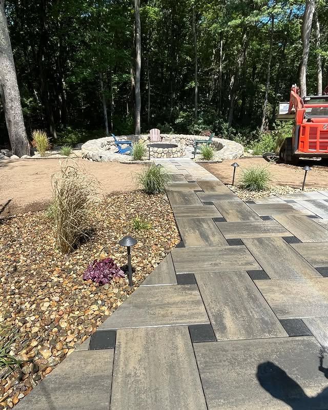 A modern paved walkway leads to a circular stone fire pit area in a wooded yard with an orange vehicle parked nearby.