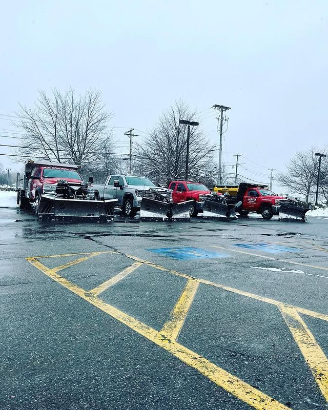 A row of pickup trucks equipped with snow plows parked in a snowy parking lot under an overcast sky.