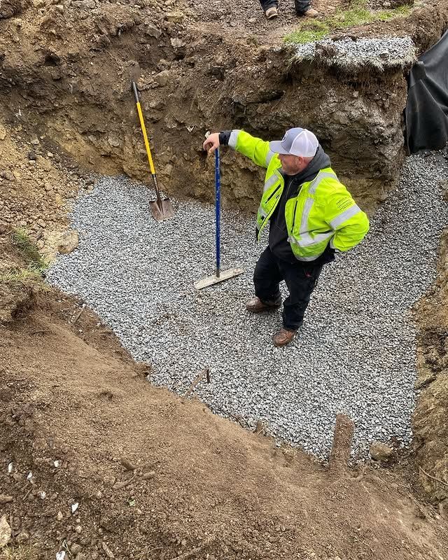 A person in a high-visibility yellow jacket stands in a gravel-filled trench with a rake and a shovel nearby.