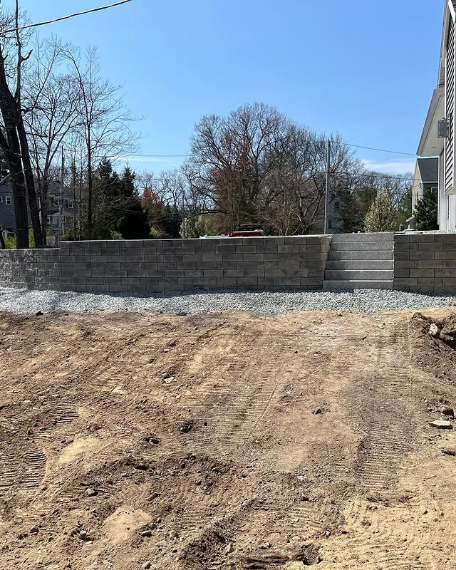 A new light gray concrete retaining wall with integrated steps in a sunny, undeveloped yard next to a house.