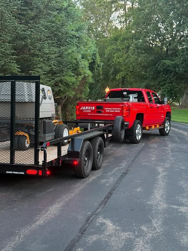 A red pickup truck is parked on an asphalt driveway, pulling a flatbed utility trailer loaded with yellow lawn equipment.