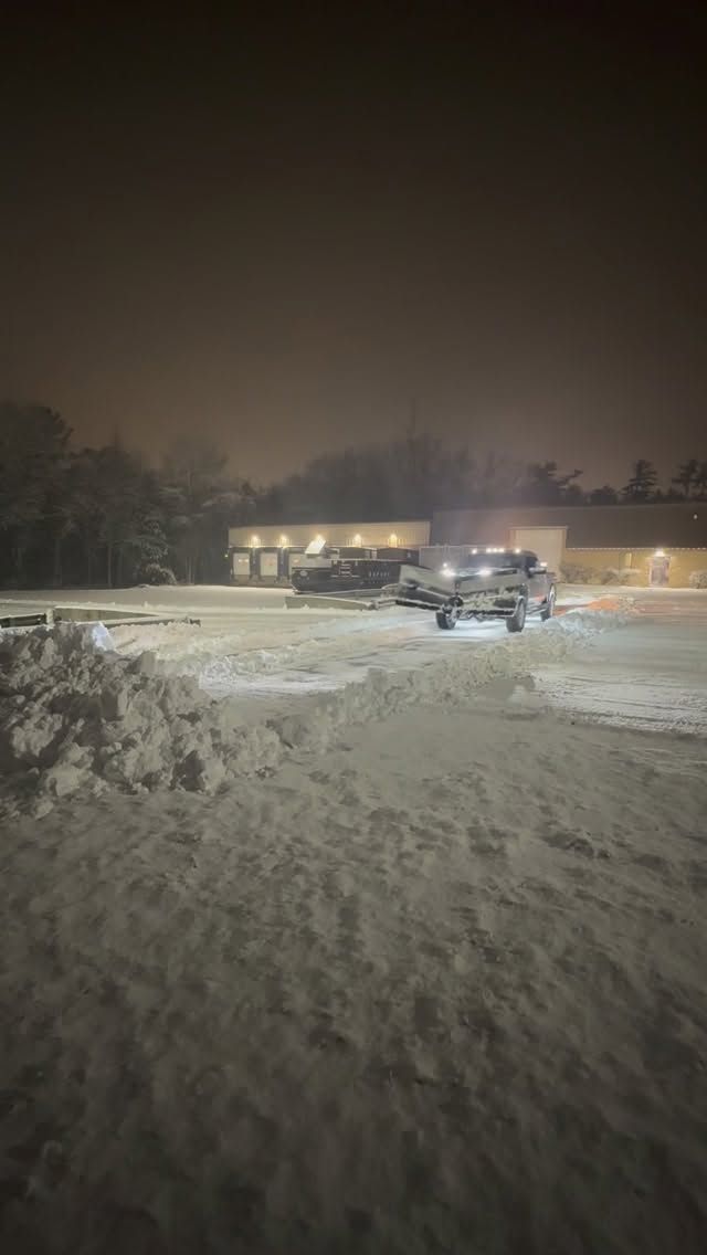 At night, a vehicle with headlights on sits in a snow-covered parking lot in front of a building.