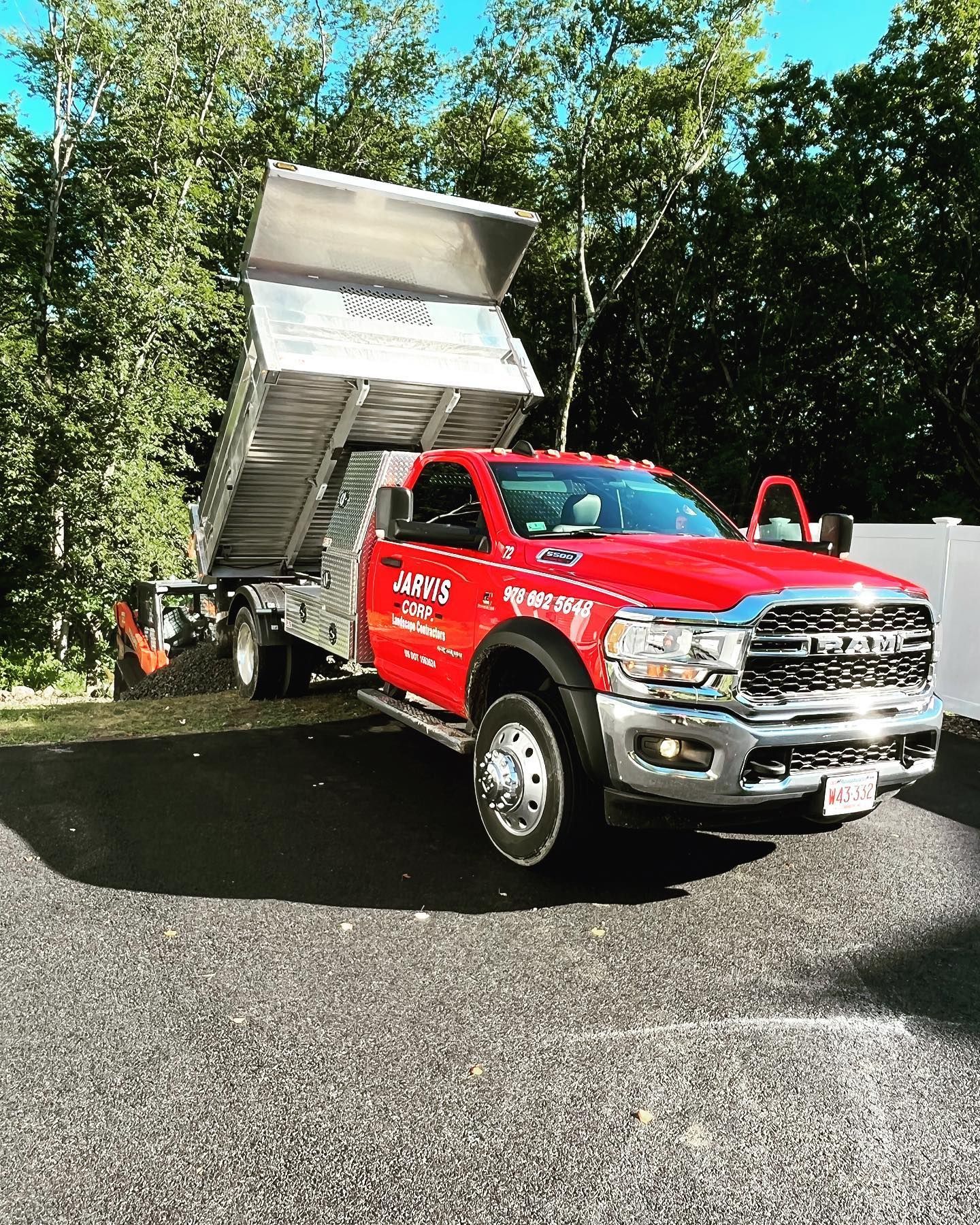 A red Ram dump truck with its bed raised, parked on a gravel surface with trees in the background under a blue sky.