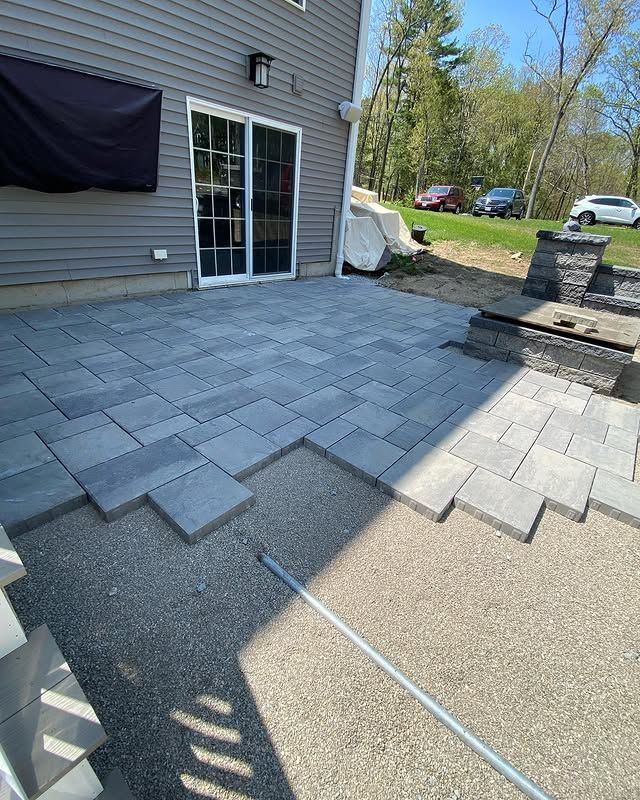 A gray paver patio under construction, partially laid over a gravel base beside a house with a sliding door and fire pit.