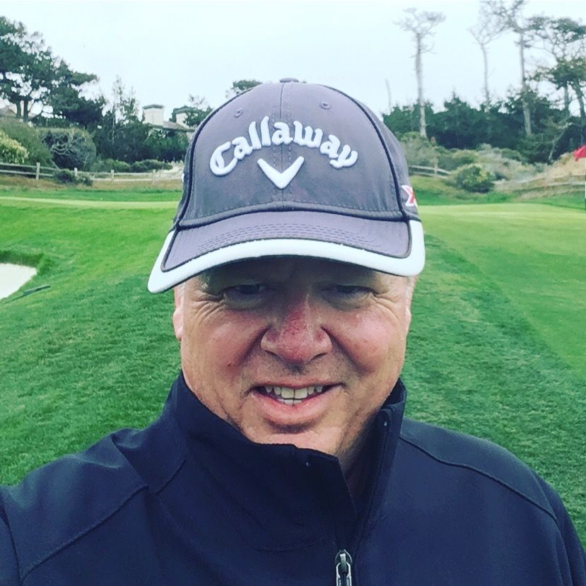 Man in gray Callaway hat smiles on a golf course, wearing a dark jacket, with trees and green grass in the background.