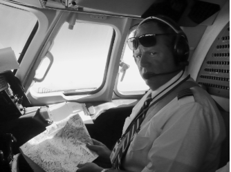 Pilot in cockpit wearing sunglasses and headset, reviewing a map. Black and white.
