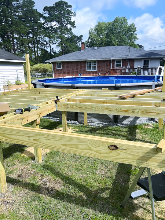 A deck being built around an above-ground pool in a backyard. Green-treated wood beams are visible, along with the pool's blue water.