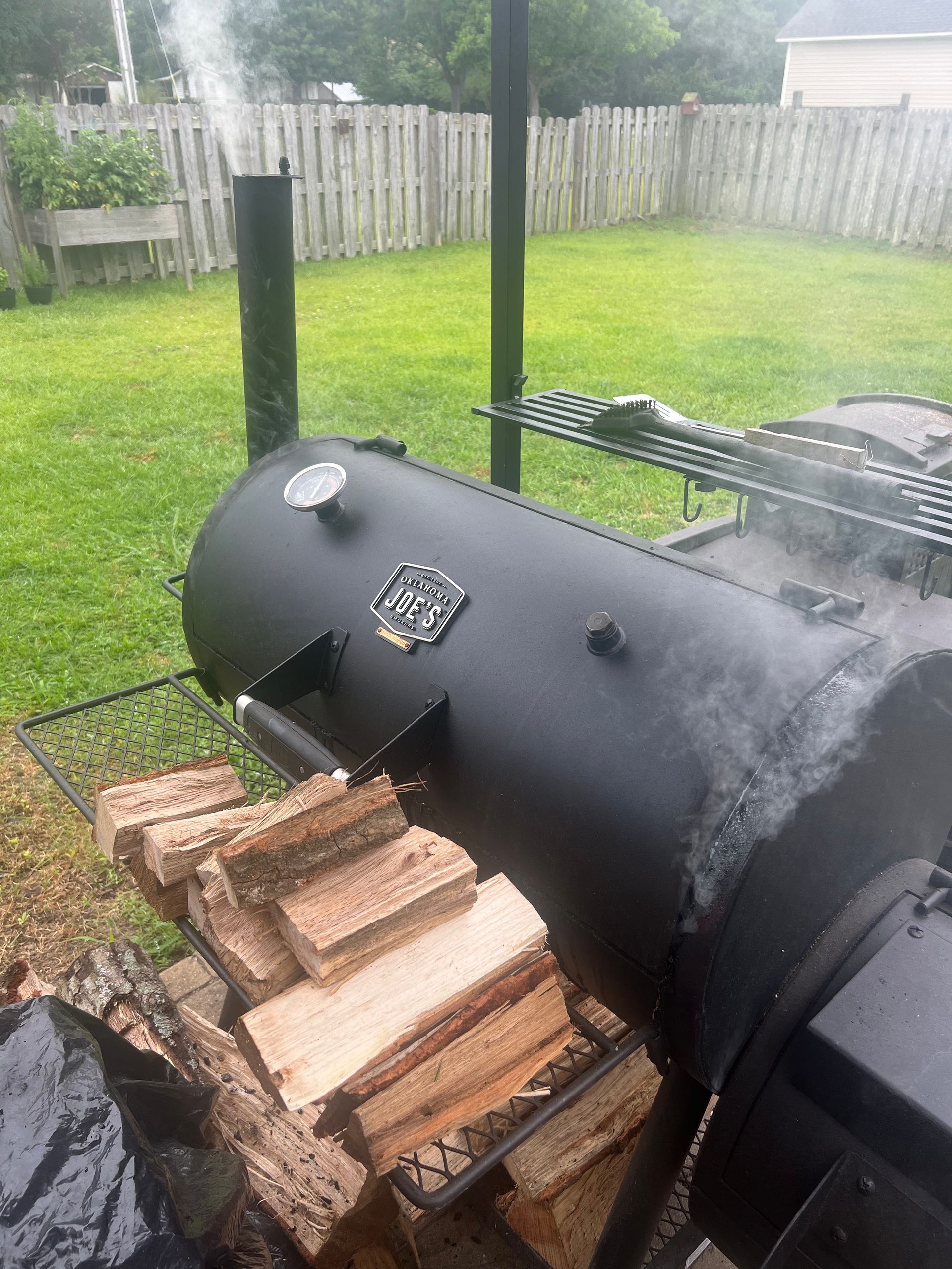 Black smoker with visible smoke plumes and firewood nearby, set in a grassy backyard.