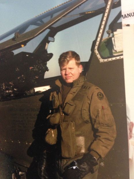 A man in a green flight suit stands next to a helicopter cockpit. He has fair skin and short blond hair.