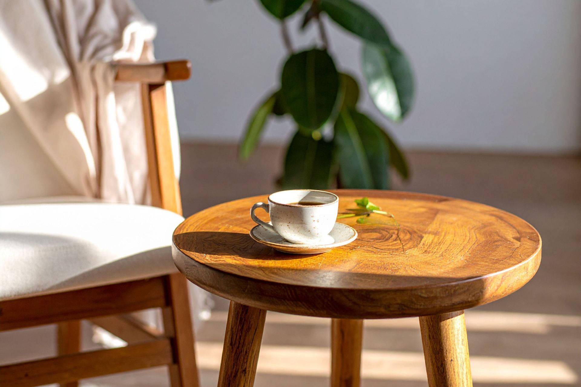 Coffee cup on a wooden stool beside a chair with a draped blanket, in front of a leafy plant.