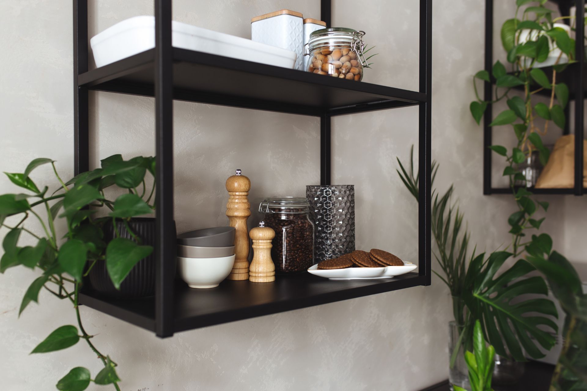 Black metal shelves with plants, jars, and kitchen items against a gray wall.