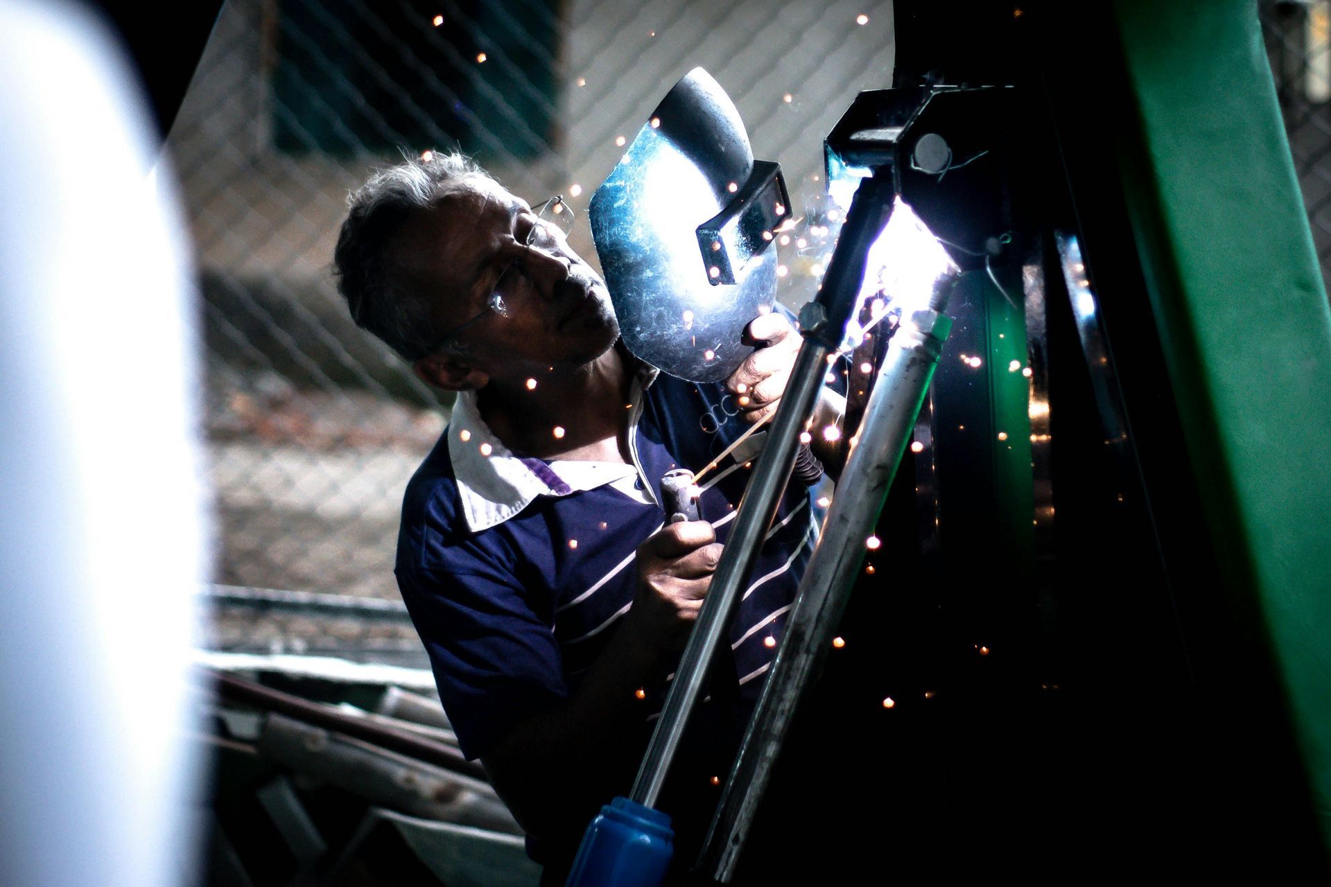 Welder in blue shirt wearing a helmet, working on metal with sparks flying.