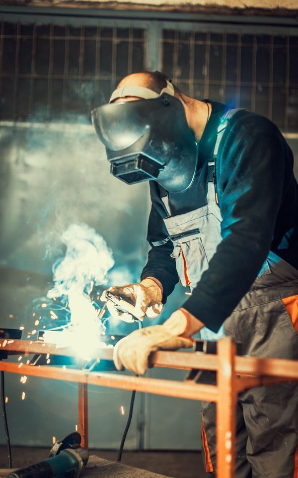 Welder in a workshop wearing a mask and gloves, working on metal with sparks flying.