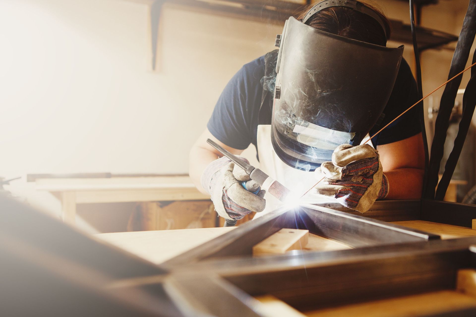 Welder wearing a mask, gloves, and protective gear working on a metal frame, creating sparks in a workshop.