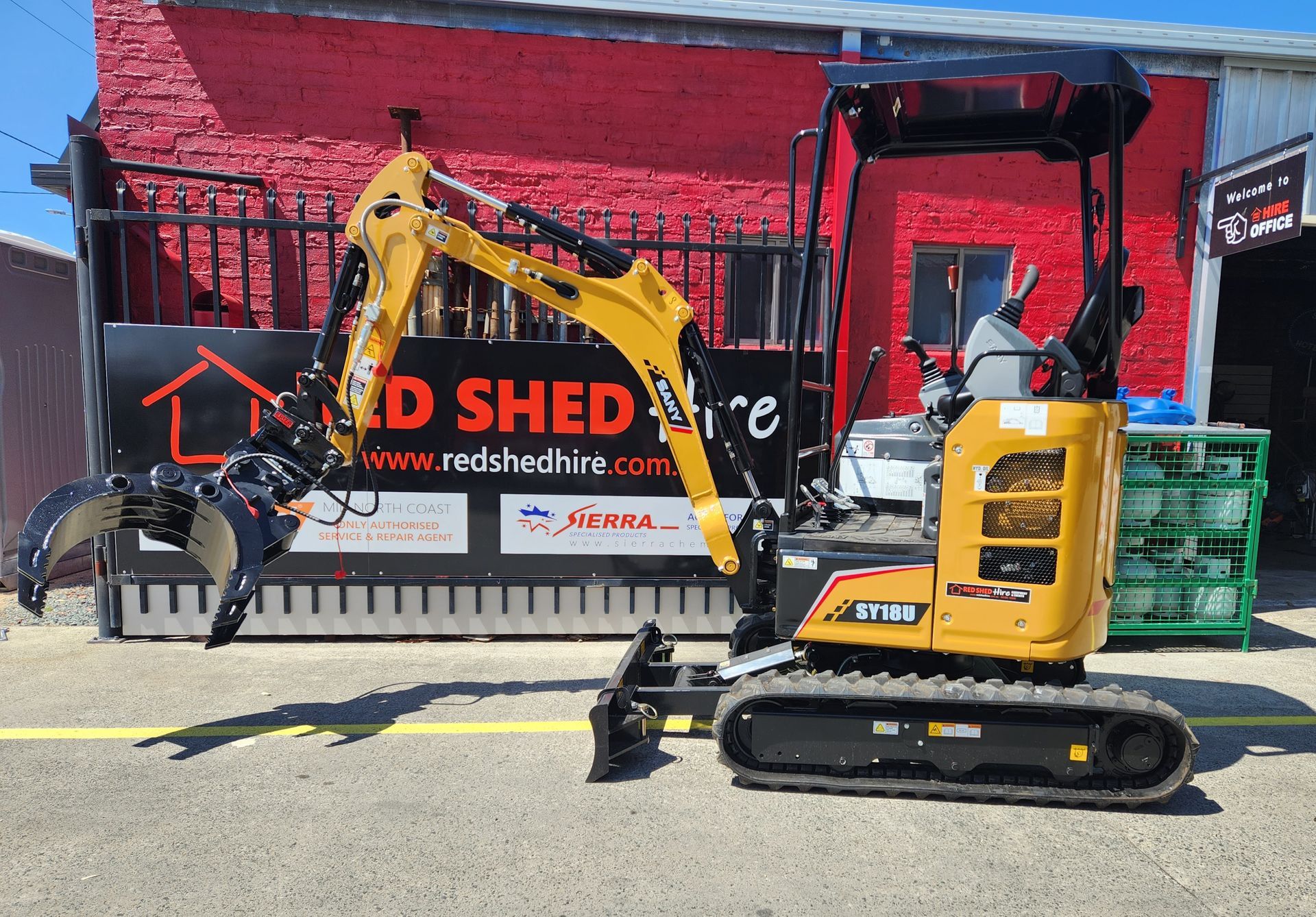 A Small Yellow Excavator Is Parked In Front Of A Red Shed — Red Shed Hire In Wauchope, NSW